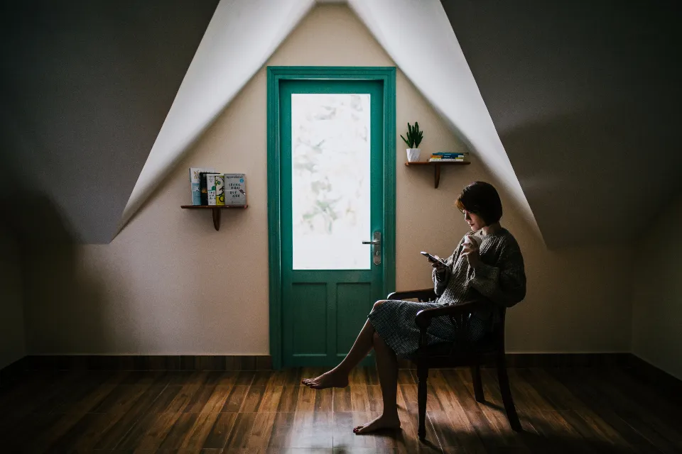 Persona sentada en una silla junto a una puerta verde, mirando el móvil en una habitación abuhardillada